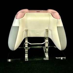 May include: A white and pink video game controller with gray grips, displayed on a clear acrylic stand. The controller has a sleek design with a matte finish. The stand is rectangular and has a modern aesthetic.