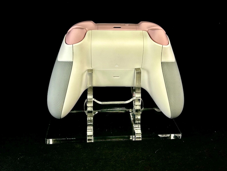 May include: A white and pink video game controller with gray grips, displayed on a clear acrylic stand. The controller has a sleek design with a matte finish. The stand is rectangular and has a modern aesthetic.