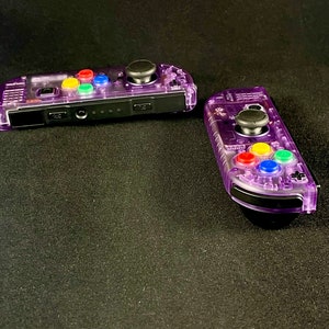 Pu&ograve; includere: Due controller Nintendo Switch Joy-Con trasparenti viola con pulsanti colorati. I controller sono su una superficie nera.