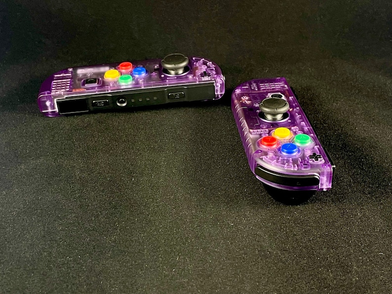 Pu&ograve; includere: Due controller Nintendo Switch Joy-Con trasparenti viola con pulsanti colorati. I controller sono su una superficie nera.