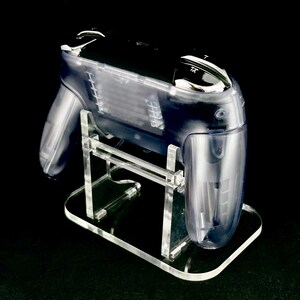 Puede incluir: Un mando de juego de pl&aacute;stico transparente y transparente sobre un soporte de acr&iacute;lico transparente. El mando tiene una parte superior negra y detalles plateados. El soporte tiene una base rectangular y sujeta el mando en &aacute;ngulo. El fondo es negro.