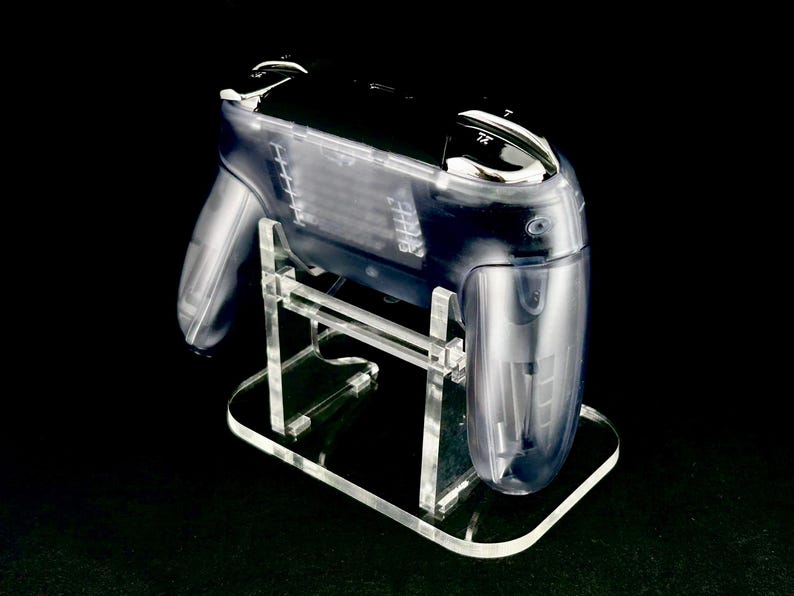 Puede incluir: Un mando de juego de pl&aacute;stico transparente y transparente sobre un soporte de acr&iacute;lico transparente. El mando tiene una parte superior negra y detalles plateados. El soporte tiene una base rectangular y sujeta el mando en &aacute;ngulo. El fondo es negro.