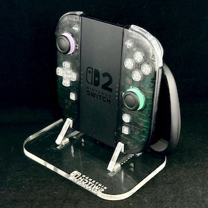 May include: A black Nintendo Switch console with transparent dark grey controller housings, featuring a purple left joystick and a teal right joystick, resting on a clear acrylic display stand with the Nintendo Switch logo etched on the base.