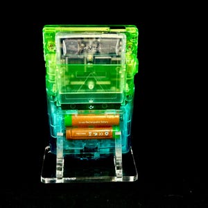 May include: A transparent, multi-layered electronic device with green and blue accents. Two orange Li-ion rechargeable batteries are visible inside. The device is mounted on a clear stand against a black background.