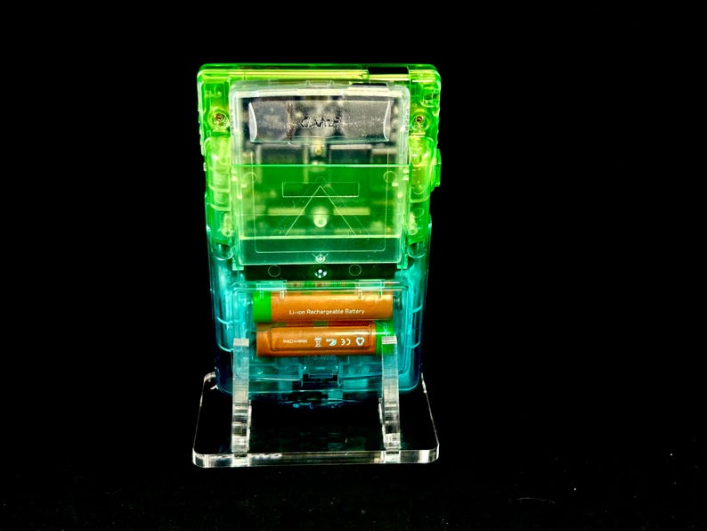 May include: A transparent, multi-layered electronic device with green and blue accents. Two orange Li-ion rechargeable batteries are visible inside. The device is mounted on a clear stand against a black background.