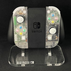 May include: A clear, transparent Nintendo Switch console with a black front panel and the text "NINTENDO SWITCH" printed on it. The console is on a clear acrylic stand with the text "NINTENDO SWITCH" printed on it.