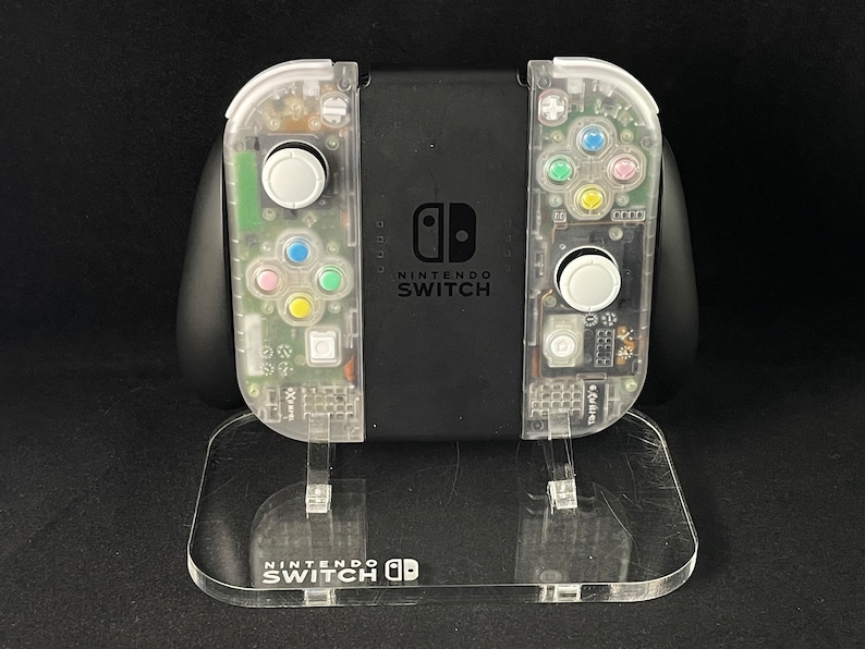 May include: A clear, transparent Nintendo Switch console with a black front panel and the text "NINTENDO SWITCH" printed on it. The console is on a clear acrylic stand with the text "NINTENDO SWITCH" printed on it.