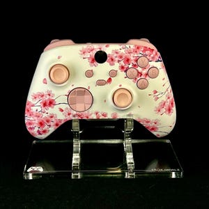 May include: White and pink Xbox Series X controller with a cherry blossom design. The controller has pink buttons and accents, and is displayed on a clear acrylic stand. The stand has the text "XBOX Series X".