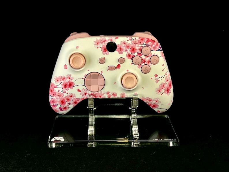 May include: White and pink Xbox Series X controller with a cherry blossom design. The controller has pink buttons and accents, and is displayed on a clear acrylic stand. The stand has the text "XBOX Series X".