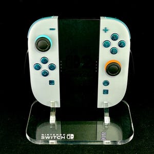 May include: A white and teal Nintendo Switch Joy-Con controller set with black joysticks and teal buttons. The controllers are displayed on a clear acrylic stand with the Nintendo Switch logo.