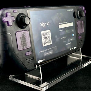 Könnte beinhalten: Eine schwarze Handheld-Spielkonsole mit violetten Knöpfen und einem Bildschirm, der eine Anmeldeseite mit einem QR-Code anzeigt. Die Konsole steht auf einem durchsichtigen Acryl-Ständer.