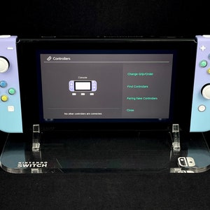May include: A purple and blue Nintendo Switch console with a clear acrylic stand. The console is displaying the "Controllers" menu on the screen, which includes options to change the grip order, find controllers, pair new controllers, and close.