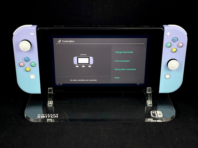 May include: A purple and blue Nintendo Switch console with a clear acrylic stand. The console is displaying the "Controllers" menu on the screen, which includes options to change the grip order, find controllers, pair new controllers, and close.