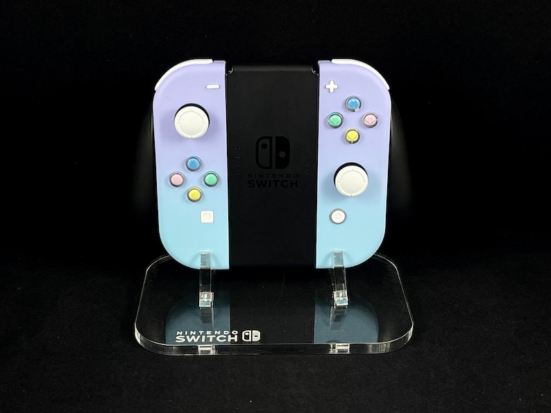 May include: A lavender and light blue Nintendo Switch console with pastel colored buttons and a clear acrylic stand. The stand has the Nintendo Switch logo printed on it.