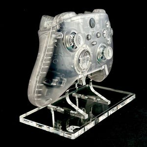 May include: A clear plastic Xbox controller on a clear acrylic stand. The controller has a textured surface and is designed for gaming.