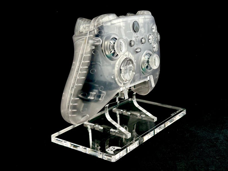 May include: A clear plastic Xbox controller on a clear acrylic stand. The controller has a textured surface and is designed for gaming.