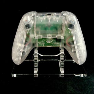 May include: A clear plastic video game controller stand with a clear plastic video game controller on top of it.
