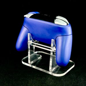 K&ouml;nnte beinhalten: Ein blauer Gamecontroller steht auf einem transparenten Acrylst&auml;nder. Der Controller hat wei&szlig;e Akzente und eine schwarze Mitte. Der St&auml;nder ist rechteckig mit einer geschwungenen Basis.