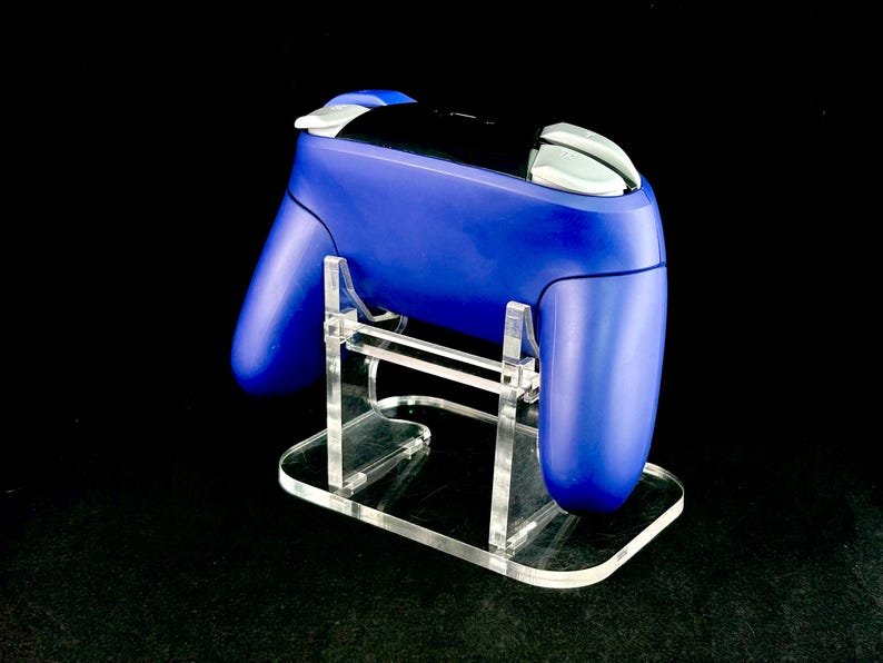 K&ouml;nnte beinhalten: Ein blauer Gamecontroller steht auf einem transparenten Acrylst&auml;nder. Der Controller hat wei&szlig;e Akzente und eine schwarze Mitte. Der St&auml;nder ist rechteckig mit einer geschwungenen Basis.