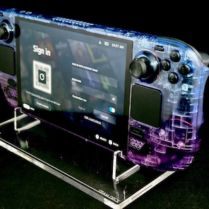 May include: A transparent blue and purple Steam Deck handheld gaming console with a screen displaying a sign-in page. The console is mounted on a clear acrylic stand. The time 10:57 AM is visible on the screen.