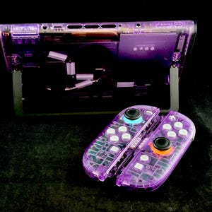 以下が含まれることがあります： 半透明の紫色のプラスチックシェルを備えた携帯型ゲーム機と、取り外し可能なコントローラーのペア。ケースを通して内部コンポーネントが見え、白いボタン、黒いジョイスティック、明るいオレンジ色のジョイスティックリングが特徴です。