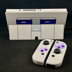 Puede incluir: Una consola Nintendo Switch blanca con un diseño de Super Nintendo Entertainment System (SNES). La consola tiene una ranura para cartuchos gris con las palabras "POWER", "RESET" y "EJECT" impresas en ella. Un mando Nintendo Switch Joy-Con blanco y gris con botones morados y blancos se muestra en primer plano.