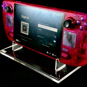 Könnte beinhalten: Eine tragbare Spielkonsole mit einem durchscheinenden pinkfarbenen Gehäuse, schwarzen Joysticks und violetten Tasten, die auf einem klaren Acrylständer steht. Der Bildschirm zeigt eine Anmeldeoberfläche mit einem QR-Code und dem Steam Deck Logo.