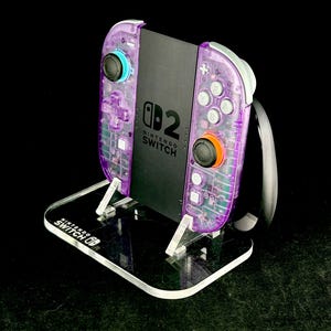 Könnte beinhalten: Eine Nintendo Switch-Konsole mit transparent-lila Joy-Con-Controllern und einem schwarzen Bildschirm, präsentiert auf einem transparenten Acryl-Ständer. Der Ständer trägt das Nintendo Switch-Logo. Die Controller haben blaue und orangefarbene Knöpfe.