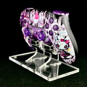Op de afbeelding: Een op maat gemaakte videogamecontroller met een uniek, artistiek ontwerp. De controller heeft een paars en wit kleurenschema met een afbeelding van een pop. Het wordt weergegeven op een heldere acrylstandaard.