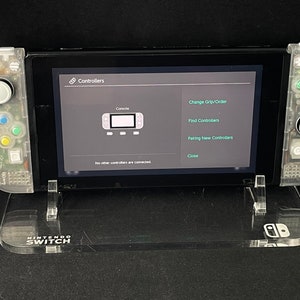 May include: A clear plastic stand with the words "NINTENDO SWITCH" printed on it. A Nintendo Switch console is sitting on the stand. The console is in a clear plastic case and the screen is showing the "Controllers" menu. The menu options include "Console", "Change Grip/Order", "Find Controllers", "Pairing New Controllers", and "Close".