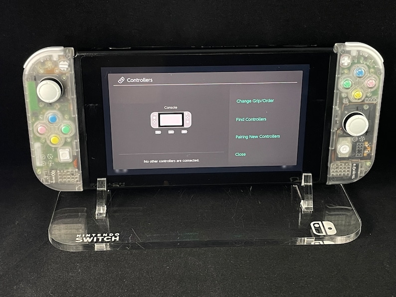 May include: A clear plastic stand with the words "NINTENDO SWITCH" printed on it. A Nintendo Switch console is sitting on the stand. The console is in a clear plastic case and the screen is showing the "Controllers" menu. The menu options include "Console", "Change Grip/Order", "Find Controllers", "Pairing New Controllers", and "Close".