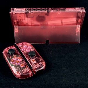 Op de afbeelding: Een transparante roze Nintendo Switch-console met bijpassende Joy-Con-controllers. De console en controllers zijn doorschijnend, waardoor de interne componenten zichtbaar zijn.