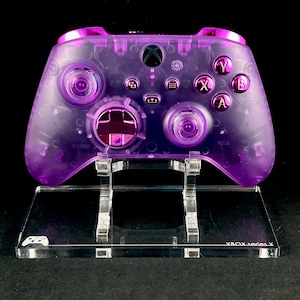 Puede incluir: Un mando Xbox Series X transparente morado con un soporte transparente morado. El mando tiene una placa frontal transparente morada con un botón Xbox transparente morado. El mando está en un soporte de acrílico transparente con el texto "XBOX Series X" impreso en la parte delantera.