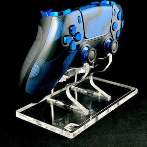 May include: A blue and black video game controller with a blue and black camouflage pattern is displayed on a clear acrylic stand.