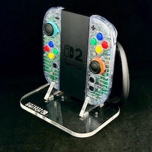 May include: A black Nintendo Switch console with clear, transparent controller housings featuring internal green circuitry and multi-coloured buttons. The console rests on a clear acrylic stand with the Nintendo Switch logo printed in white on the base.
