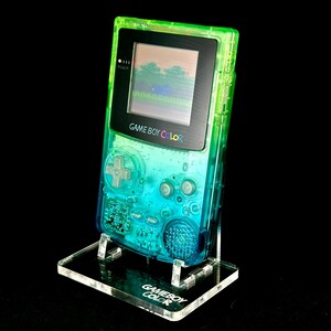 May include: A transparent Game Boy Color handheld gaming console with a gradient green and blue design. The screen displays a pixelated landscape. The console is on a clear acrylic stand with the words "GAME BOY COLOR" printed on it.