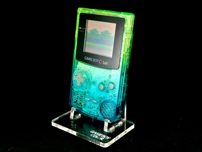 May include: A transparent Game Boy Color handheld gaming console with a gradient green and blue design. The screen displays a pixelated landscape. The console is on a clear acrylic stand with the words "GAME BOY COLOR" printed on it.