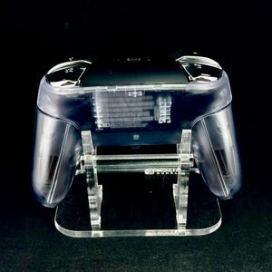Puede incluir: Un mando de juego transparente sobre un soporte de acr&iacute;lico transparente. Los componentes internos del mando son visibles. El soporte tiene una base rectangular y sostiene el mando con soportes verticales.