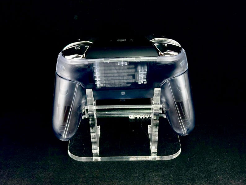 Puede incluir: Un mando de juego transparente sobre un soporte de acr&iacute;lico transparente. Los componentes internos del mando son visibles. El soporte tiene una base rectangular y sostiene el mando con soportes verticales.
