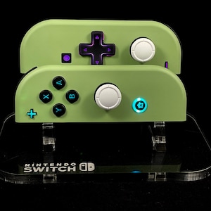 May include: Two green Nintendo Switch controllers with a clear plastic stand. The stand has the words "Nintendo Switch" printed on it.