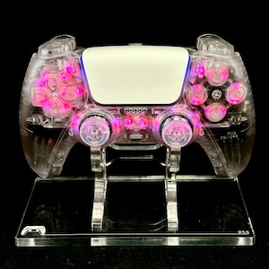 May include: A clear, transparent gaming controller with pink and blue internal lighting, displayed on a clear acrylic stand. The controller has a white center panel and pink buttons. The stand has a small controller icon and the text "PS5".