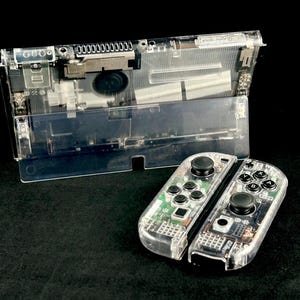 May include: Clear plastic video game console with visible internal components. Two clear controllers with black buttons are in the foreground. The console is open, revealing its internal structure. The background is black.