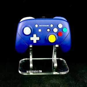 K&ouml;nnte beinhalten: Ein blauer SwitchHouse-Controller mit wei&szlig;en, roten, gr&uuml;nen und gelben Kn&ouml;pfen, pr&auml;sentiert auf einem transparenten Acrylst&auml;nder. Der St&auml;nder hat das Nintendo Switch-Logo. Der Controller hat ein wei&szlig;es kreuzf&ouml;rmiges Steuerkreuz.