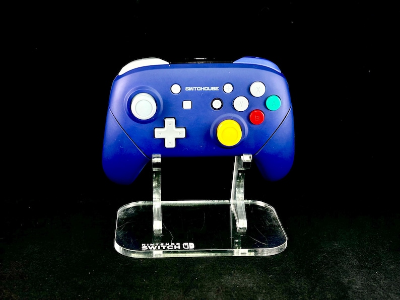 K&ouml;nnte beinhalten: Ein blauer SwitchHouse-Controller mit wei&szlig;en, roten, gr&uuml;nen und gelben Kn&ouml;pfen, pr&auml;sentiert auf einem transparenten Acrylst&auml;nder. Der St&auml;nder hat das Nintendo Switch-Logo. Der Controller hat ein wei&szlig;es kreuzf&ouml;rmiges Steuerkreuz.