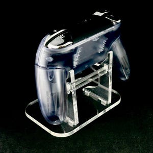 Puede incluir: Un soporte transparente de acr&iacute;lico para mando de videojuegos que sostiene un mando transparente. El soporte tiene una base rectangular y sujeta el mando en &aacute;ngulo. El mando tiene botones negros y un dise&ntilde;o elegante, sobre un fondo negro.
