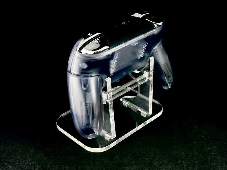 Puede incluir: Un soporte transparente de acr&iacute;lico para mando de videojuegos que sostiene un mando transparente. El soporte tiene una base rectangular y sujeta el mando en &aacute;ngulo. El mando tiene botones negros y un dise&ntilde;o elegante, sobre un fondo negro.