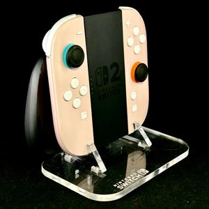 May include: A Nintendo Switch console with pink Joy-Con controllers, black joysticks, and white buttons, displayed on a clear acrylic stand. The stand has the Nintendo Switch logo. The console's screen displays the Nintendo Switch logo.