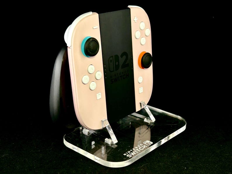 May include: A Nintendo Switch console with pink Joy-Con controllers, black joysticks, and white buttons, displayed on a clear acrylic stand. The stand has the Nintendo Switch logo. The console's screen displays the Nintendo Switch logo.