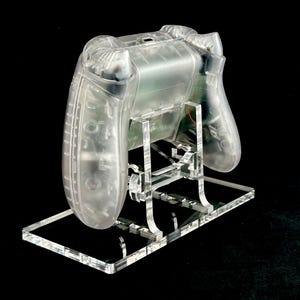 May include: A clear plastic video game controller stand. The stand is made of acrylic and has a clear, transparent finish. The stand is designed to hold a video game controller in a vertical position.