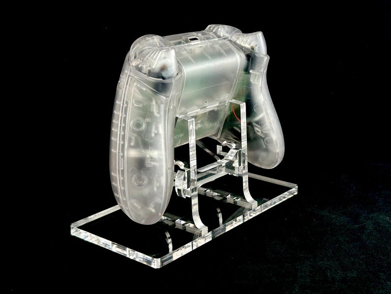 May include: A clear plastic video game controller stand. The stand is made of acrylic and has a clear, transparent finish. The stand is designed to hold a video game controller in a vertical position.